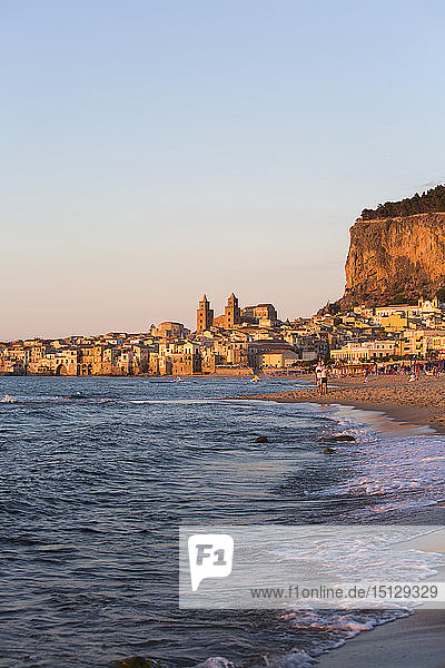 Blick vom Strand entlang der Wasserkante auf die Stadt und die zum UNESCO-Weltkulturerbe gehörende arabisch-normannische Kathedrale  Sonnenuntergang  Cefalu  Palermo  Sizilien  Italien  Mittelmeer  Europa