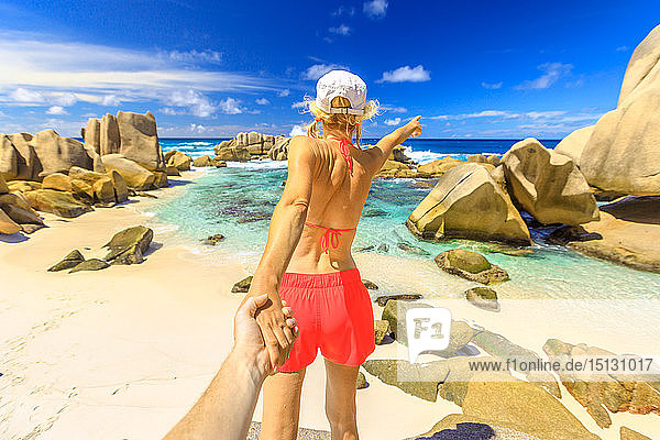 Blonde Touristin im orangefarbenen Bikini hält die Hand ihres Partners an der Anse Marron  Insel La Digue  Seychellen  Indischer Ozean  Afrika