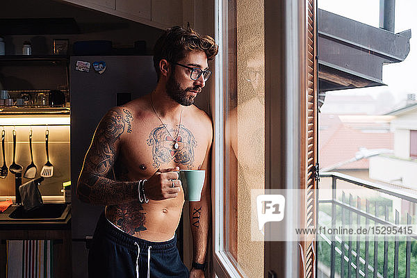 Mittelgroßer erwachsener Mann mit Tätowierungen  der durch ein Küchenfenster hinaussieht