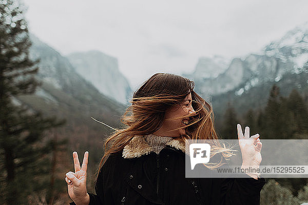 Junge Frau  die lange braune Haare schüttelt und ein Friedenszeichen in einer Berglandschaft setzt  Yosemite Village  Kalifornien  USA
