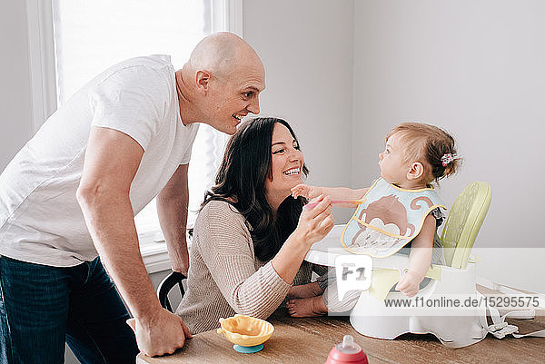 Mutter und Vater füttern ihre Tochter im Kindersitz auf dem Küchentisch
