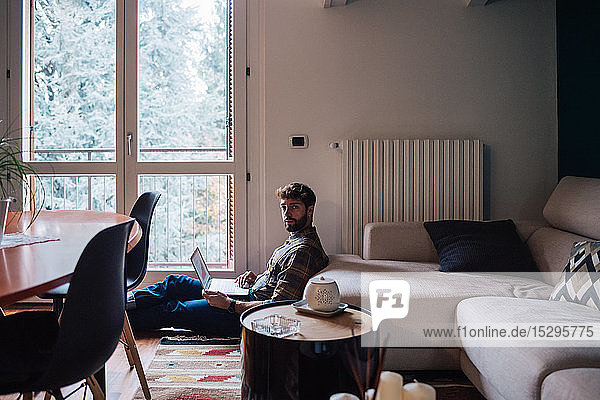Mittelgroßer erwachsener Mann sitzt mit Laptop auf dem Wohnzimmerboden  Portrait