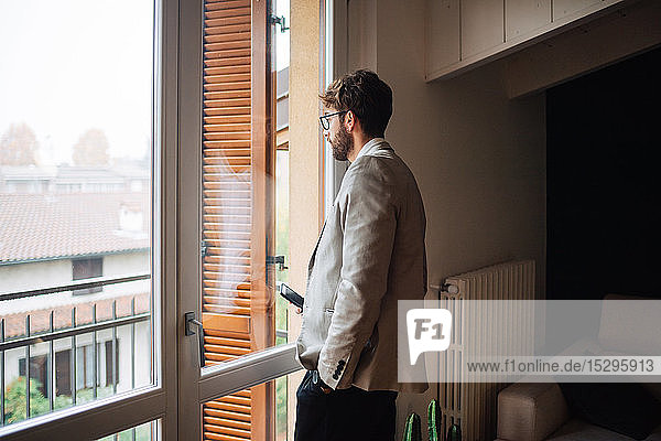 Mittelgroßer erwachsener Mann mit Smartphone in der Hand  der durch ein Wohnungsfenster hinausblickt