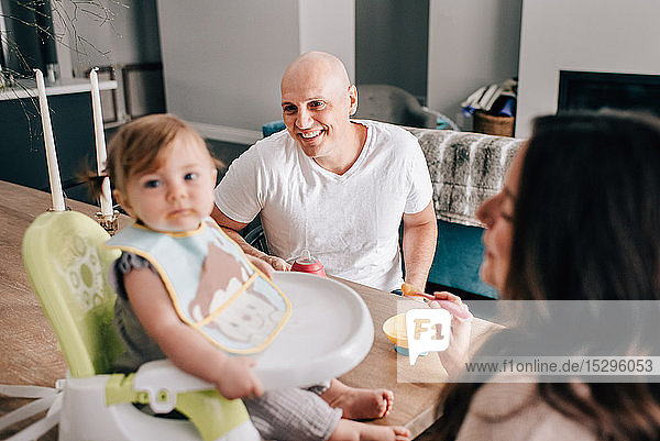 Mutter und Vater füttern ihre Tochter im Kindersitz auf dem Küchentisch