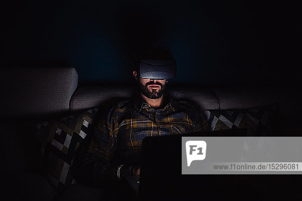 Bärtiger mittelgroßer Mann sitzt im Dunkeln auf dem Sofa und schaut durch ein Virtual-Reality-Headset