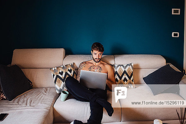 Mittelgroßer erwachsener Mann mit tätowierter Brust auf Sofa mit Laptop