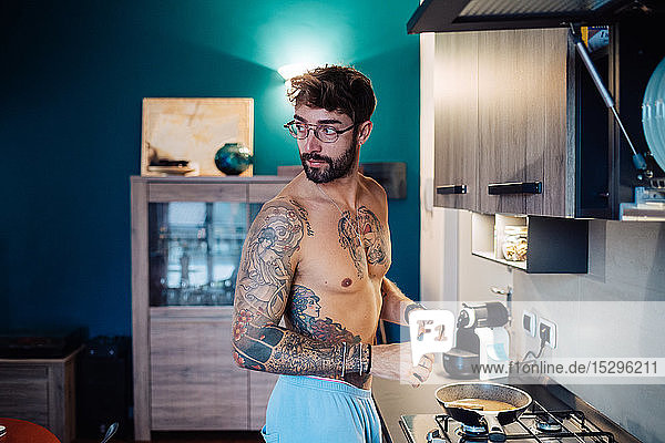 Mittelgroßer erwachsener Mann mit tätowierter Brust  der das Frühstück vorbereitet und dabei über die Schulter schaut