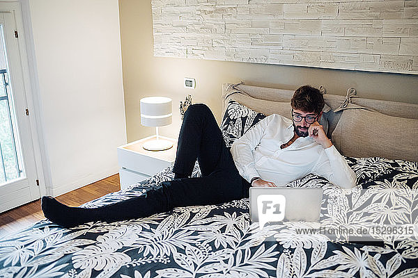 Mittelgroßer erwachsener Mann  der mit einem Laptop auf dem Bett liegt