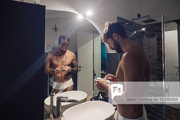 Mid adult man with tattoos using smartphone touchscreen at bathroom mirror  mirror image
