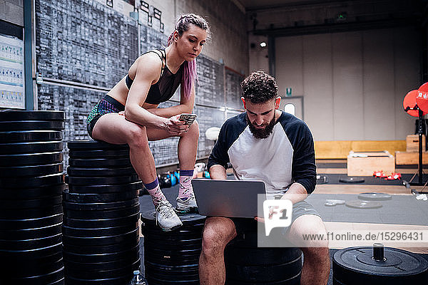Junge Frau und Mann trainieren gemeinsam im Fitnessstudio  sitzen auf Gewichten und schauen auf den Laptop