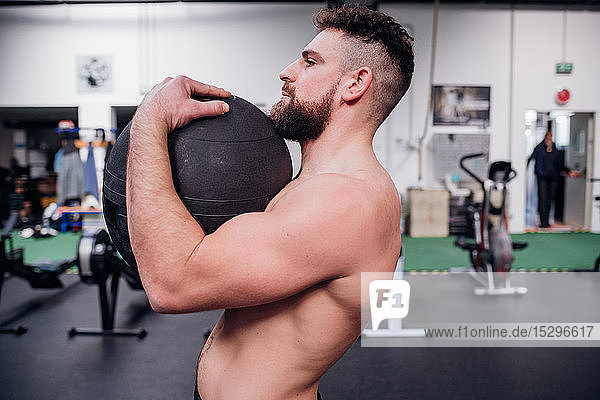 Young man training  lifting atlas ball in gym  side view