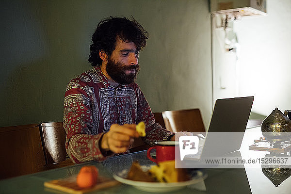 Bärtiger junger Mann frühstückt mit Laptop zu Hause