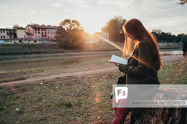 Young woman with long red hair sitting on tree stump reading book on riverside at sunset  Florence  Tuscany  Italy