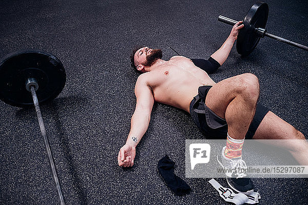 Nacktbrust trainierender junger Mann  der erschöpft zwischen Hanteln in der Turnhalle liegt