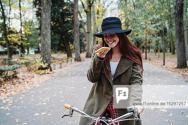 Junge Frau mit langen roten Haaren auf Fahrrad schaut auf Smartphone im Herbstpark  Florenz  Toskana  Italien