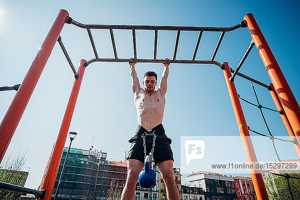 Gymnastik in der Turnhalle im Freien  junger Mann mit nackter Brust  der mit einer Kettlebell am Hüftgurt an einem Übungsgerät hängt