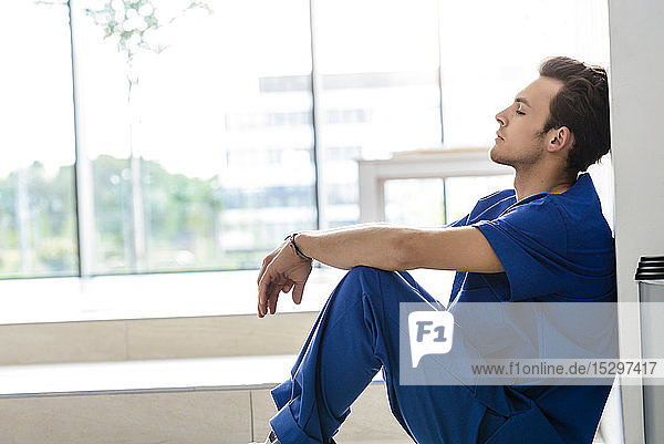 Junger männlicher Assistenzarzt sitzt auf dem Boden im Krankenhauskorridor