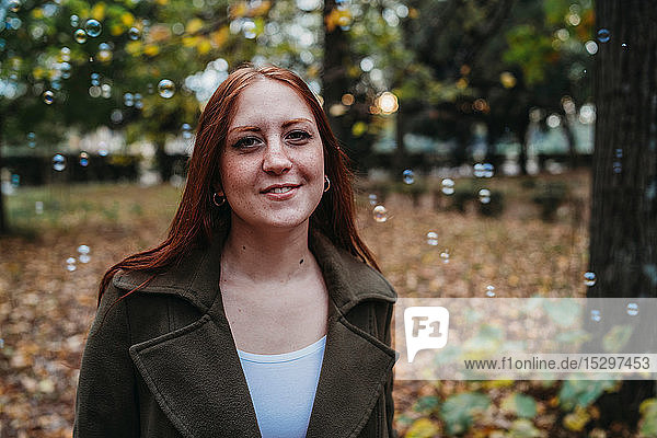 Junge Frau mit langen roten Haaren zwischen schwebenden Blasen im Herbstpark  Kopf- und Schulterporträt