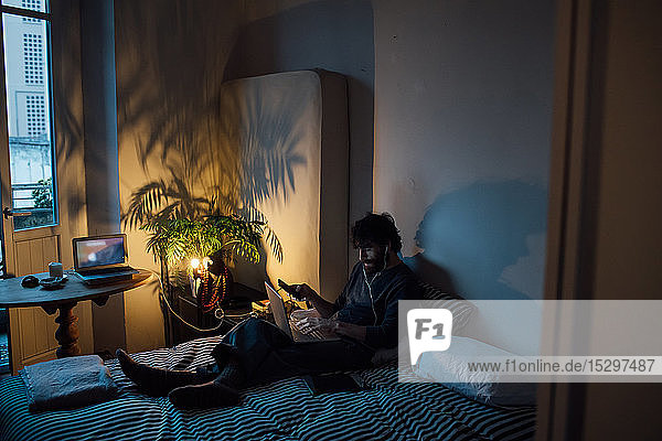 Bärtiger junger Mann mit Smartphone und Laptop im Bett