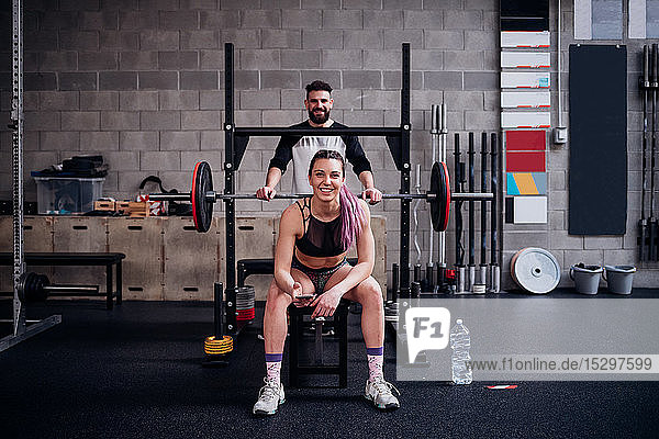 Junge Frau und Mann trainieren gemeinsam im Fitnessstudio  sitzen auf Hantelbank  Porträt
