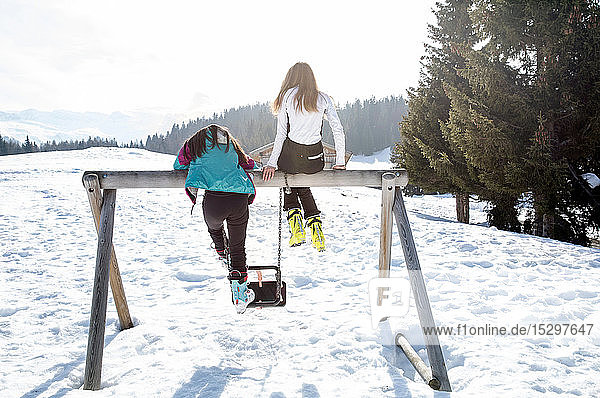 Zwei jugendliche Skifahrerinnen auf der Schaukel in schneebedeckter Landschaft  Rückansicht  Tirol  Steiermark  Österreich