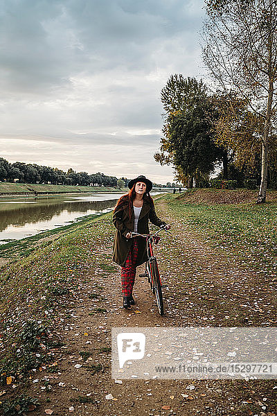 Junge Frau mit langen roten Haaren  die am Flussufer ein Fahrrad schiebt  Porträt in voller Länge  Florenz  Toskana  Italien