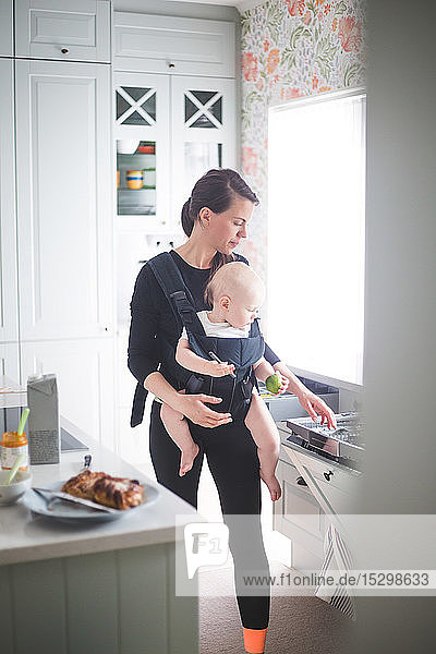 Mother carrying daughter in baby carrier while standing in kitchen at home