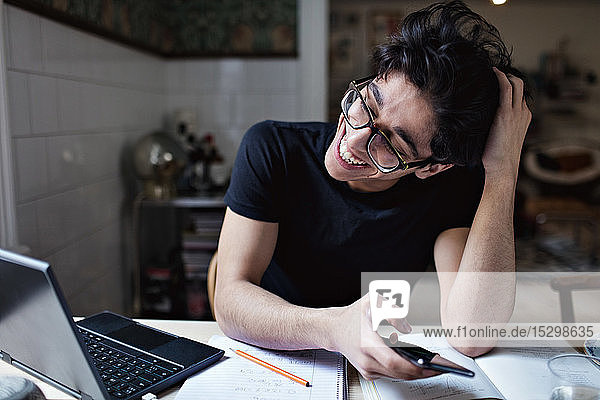 Fröhlicher  nach sozialen Medien süchtiger junger Mann benutzt Mobiltelefon während er Hausaufgaben macht