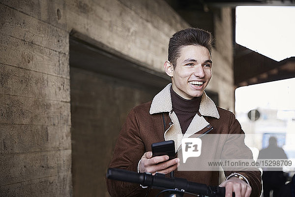 Lächelnder Teenager mit elektrischem Schubsroller schaut weg  während er sein Mobiltelefon unter der Brücke hält