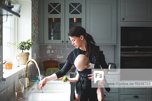 Mutter wäscht Hand in Küchenspüle  während sie ein Mädchen zu Hause trägt