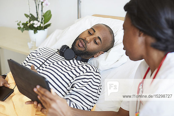 Männlicher Patient betrachtet digitales Tablett in der Hand einer Ärztin auf der Krankenhausstation