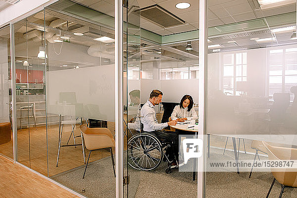 Geschäftsmann sitzt im Rollstuhl  während er mit einer Kollegin im Sitzungssaal am Arbeitsplatz mit Blick durch die Tür arbeitet