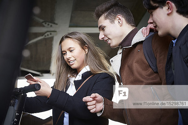 Teenage boys looking at female friend using mobile phone below bridge