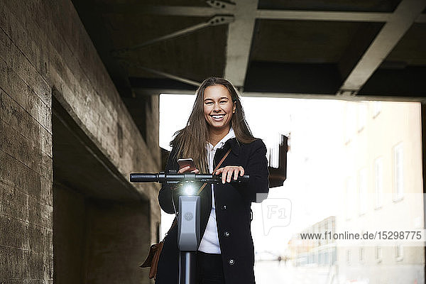 Porträt eines lächelnden Teenager-Mädchens mit elektrischem Schieberoller  das sein Mobiltelefon unter der Brücke hält