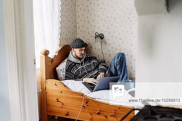 Junger Mann benutzt Laptop  während er sich auf dem Bett ausruht und durch die Tür zu Hause gesehen wird