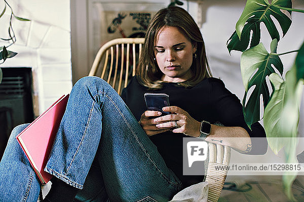 Frau sitzt mit einem Smartphone mit einem Buch auf einem Sessel im Zimmer zu Hause