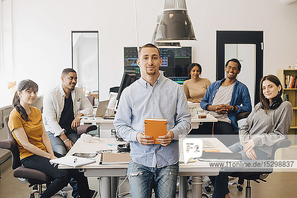 Portrait of confident businessman holding book while standing with colleagues sitting in background at creative office