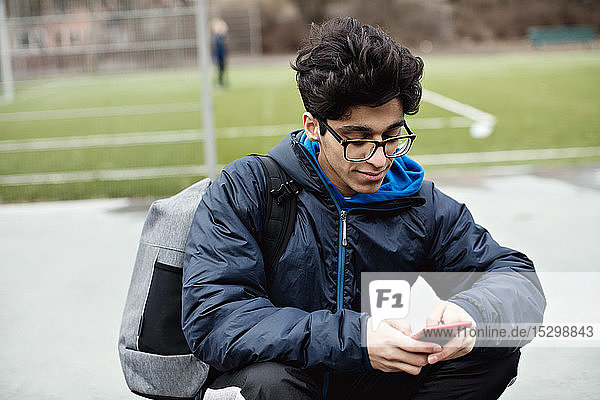 Junger Mann benutzt Mobiltelefon  während er im Winter in der Stadt gegen ein Fußballfeld sitzt