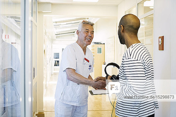 Senior doctor with clipboard greeting young man at hospital corridor
