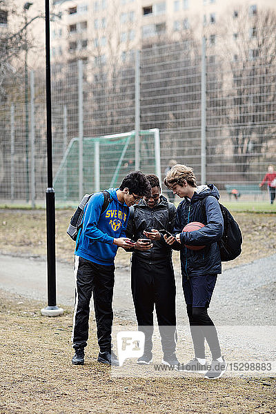 Full length of social media addicted friends wearing sports clothing while using mobile phones on playing field
