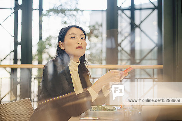 Zuversichtliche Geschäftsfrau mit Handy in der Cafeteria sitzend im Büro