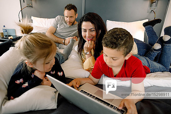 Eltern und Mädchen mit einem Jungen  der einen Laptop am Bett im Hotelzimmer benutzt