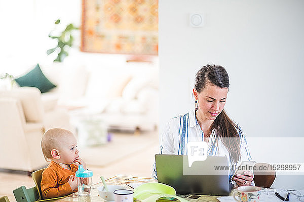 Baby girl looking at busy working mother while sitting at table in home office