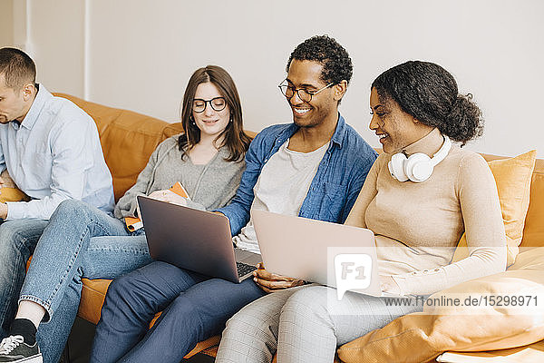 Lächelnde Programmierer  die über Laptops arbeiten  während sie im Kreativbüro auf dem Sofa sitzen