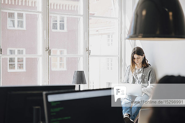 Fokussierte junge Programmiererin  die einen Laptop benutzt  während sie im Kreativbüro am Fenster sitzt
