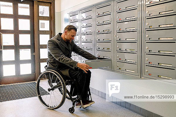 Behinderter erwachsener Mann öffnet in voller Länge den Briefkasten in der Umkleidekabine  während er auf einem Rollstuhl sitzt