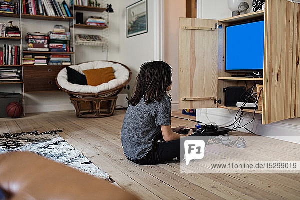 Seitenansicht eines Jungen  der ein Videospiel spielt  während er zu Hause auf einem Hartholzboden sitzt