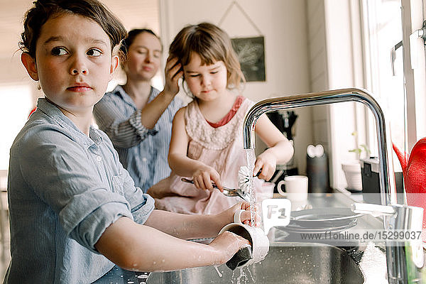 Mädchen wäscht Becher an der Küchenspüle mit Schwester und Mutter im Hintergrund zu Hause