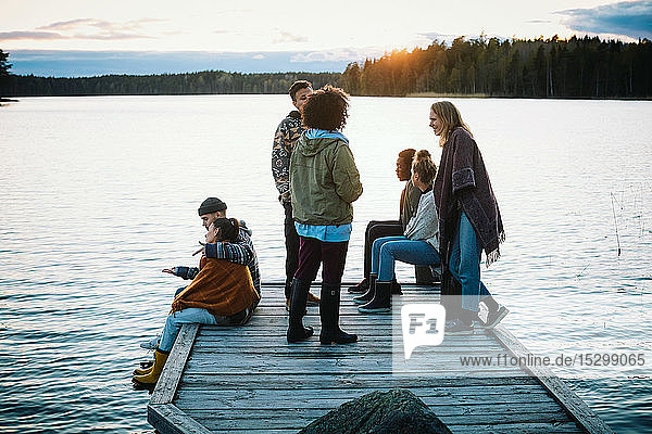 Freundinnen und Freunde unterhalten sich bei Sonnenuntergang auf der Mole über dem See gegen den Himmel