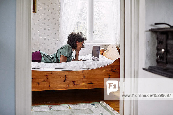 Seitenansicht einer jungen Frau  die einen Laptop benutzt  während sie zu Hause auf dem Bett liegt  durch die Tür gesehen